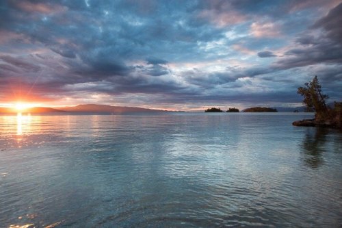 A serene lake at sunset, with vibrant clouds reflecting on the water and distant islands silhouetted against the horizon.