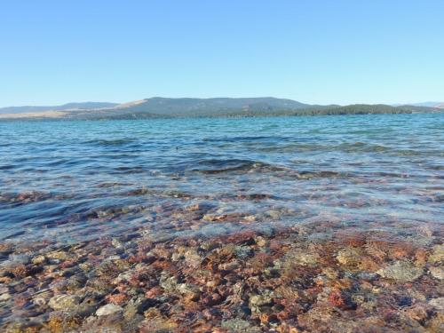 Clear water reveals colorful stones beneath the surface, with mountains in the background under a blue sky.