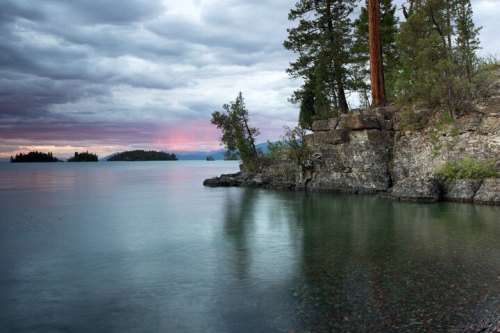 Serene lake scene with rocky shore, trees, and a cloudy sky reflecting soft colors at sunset.