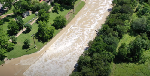 Aerial view of a flooded river surrounded by green trees and parkland, with muddy water flowing rapidly.