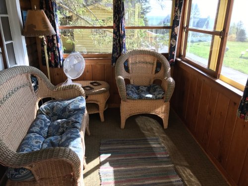 A cozy sunroom with wicker furniture, a small table, and a fan, surrounded by large windows and colorful curtains.