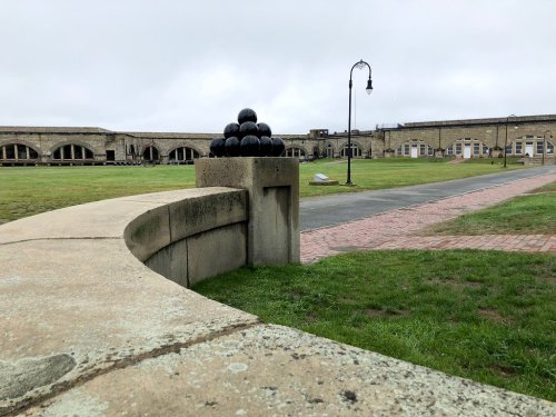 A stone wall curves around a grassy area, topped with a pyramid of black cannonballs, with historic buildings in the background.