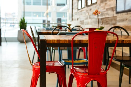 A cozy café interior featuring a wooden table and colorful metal chairs, with a small plant on the table.