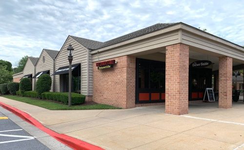 A brick and siding building with multiple storefronts, featuring a sidewalk and landscaped greenery.