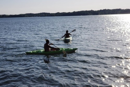 Two people kayaking on a calm lake, surrounded by trees and reflecting sunlight on the water.
