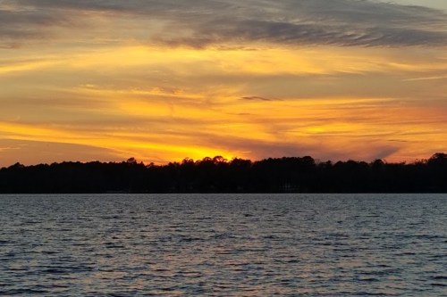 A vibrant sunset over a calm lake, with orange and yellow hues reflecting on the water and silhouetted trees in the background.