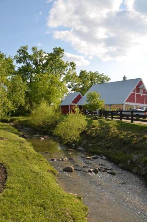 A red barn beside a stream, surrounded by green trees and grass under a blue sky with fluffy clouds.