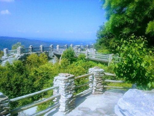 Scenic view of a stone bridge and lush greenery against a clear blue sky. Mountains visible in the distance.