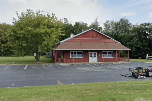 A red and brown single-story building with a porch, surrounded by trees and a grassy area, with a parking lot in front.