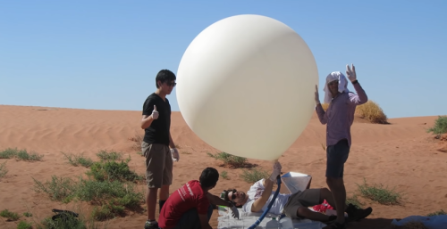 A group of people prepares a large balloon in a desert setting, with sand dunes and clear blue sky in the background.