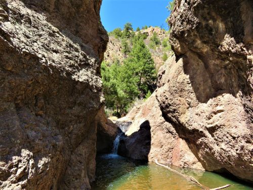 A narrow canyon with steep rock walls and a small waterfall flowing into a serene pool of water under a clear blue sky.