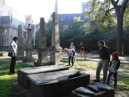 A group of people explores a historic cemetery with tall gravestones and greenery in the background.