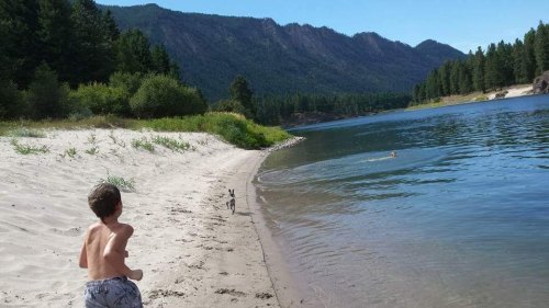 A boy runs along a sandy beach by a river, with a dog playing nearby and mountains in the background.