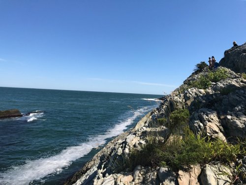 A rocky coastline under a clear blue sky, with waves gently crashing against the shore and people exploring the area.