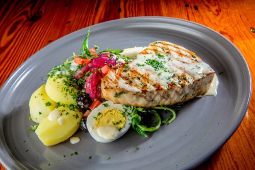 Grilled fish fillet on a plate with potatoes, salad, and a drizzle of sauce, garnished with herbs.