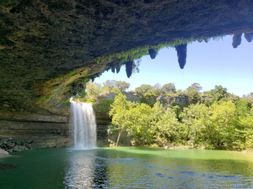 A serene waterfall cascades into a green pool, framed by rocky overhangs and lush trees under a clear blue sky.