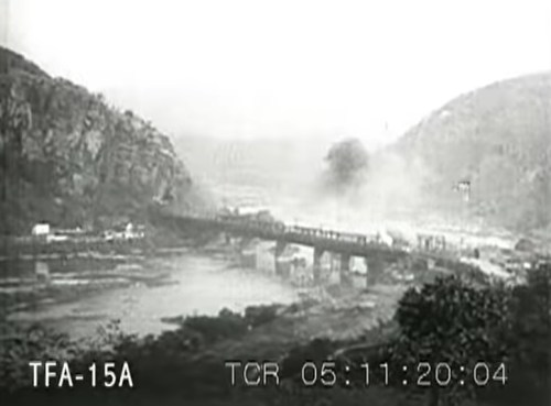 Black and white image of a bridge over a river, surrounded by misty mountains and a small settlement.