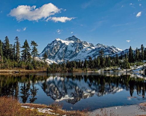A serene mountain landscape with snow-capped peaks reflecting in a calm lake, surrounded by evergreen trees and blue sky.