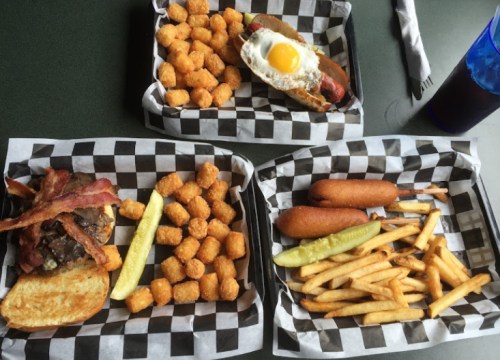 A meal featuring a burger with bacon, tater tots, a hot dog with an egg, corn dogs, and fries on checkered trays.