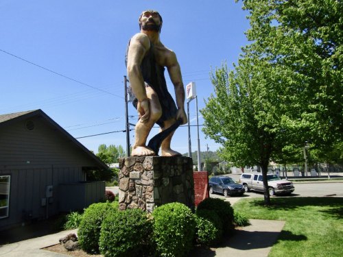 A large statue of a muscular figure stands on a stone pedestal, surrounded by greenery and a clear blue sky.