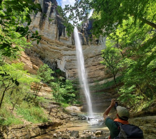 A person takes a photo of a tall waterfall surrounded by lush greenery and rocky cliffs.
