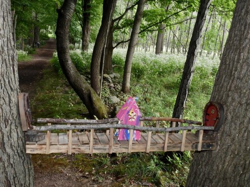 A whimsical wooden bridge between two trees, featuring a small pink tent and a tiny door, surrounded by lush greenery.