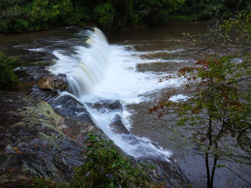 A waterfall cascades over rocks into a river, surrounded by lush greenery and autumn foliage.