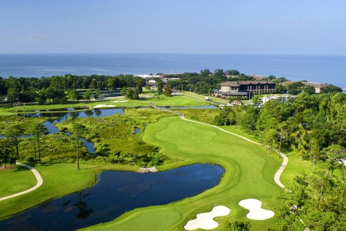 Aerial view of a golf course with water features, lush greenery, and a hotel by the ocean in the background.