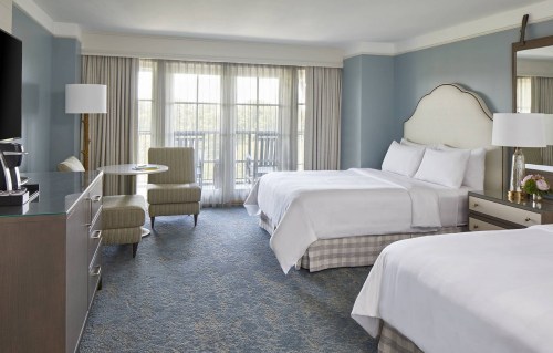 A bright hotel room featuring two beds, a seating area, a TV, and large windows with natural light.