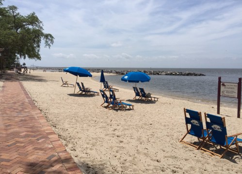 A sandy beach with blue umbrellas and chairs, lined by a brick walkway and calm water in the background.