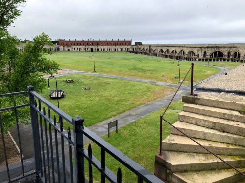 A grassy courtyard surrounded by stone buildings, viewed from a balcony with stairs in the foreground.