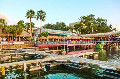 A waterfront scene featuring a tiki-style restaurant with palm trees and docks on a calm river.