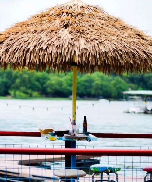 A thatched umbrella over a table with drinks, overlooking a calm lake and boats in the background.