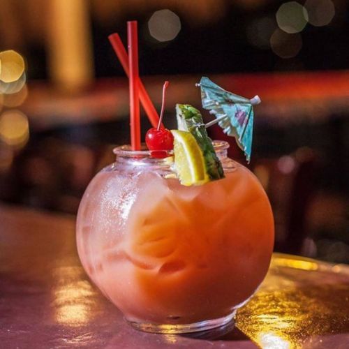 A colorful cocktail in a fishbowl glass, garnished with a cherry, lemon, and a small umbrella, on a bar surface.