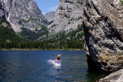 A person splashes in a clear lake surrounded by towering mountains and lush greenery.