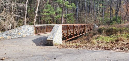 A metal bridge with a stone base spans a small creek, surrounded by trees and autumn foliage.