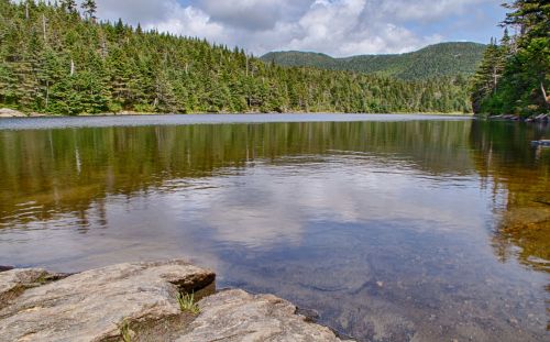 A serene lake surrounded by lush green trees and mountains under a partly cloudy sky.