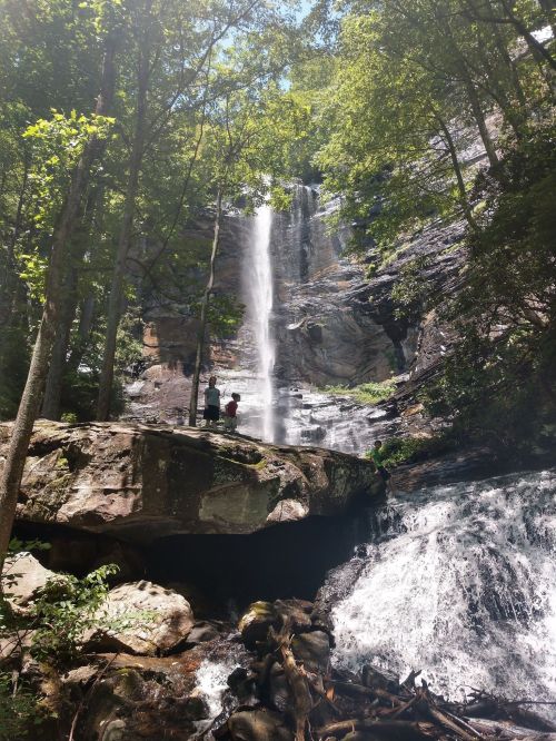 A tall waterfall cascades down rocky cliffs, surrounded by lush green trees, with two people standing on a large rock below.