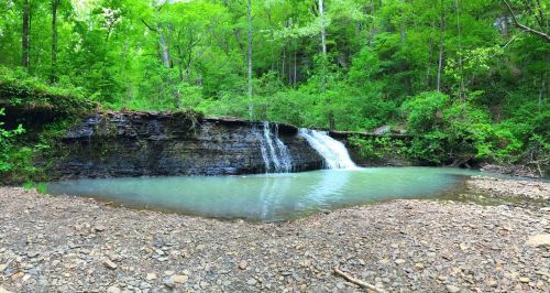 A serene waterfall cascades into a calm pool, surrounded by lush green trees and rocky terrain.