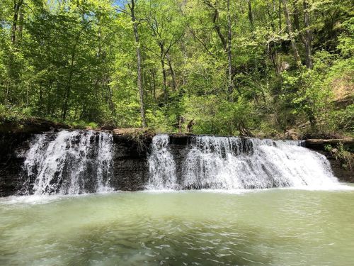 A serene waterfall cascading over rocks into a clear, greenish pool, surrounded by lush green trees.