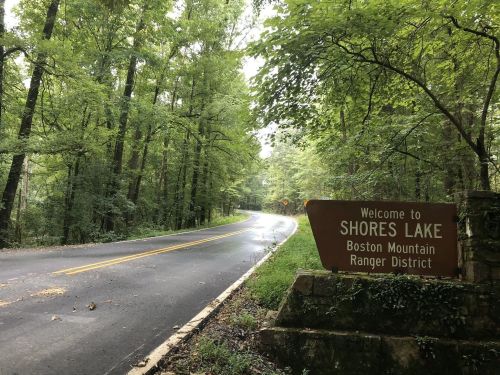 A winding road leads to a sign welcoming visitors to Shores Lake in the Boston Mountain Ranger District, surrounded by trees.
