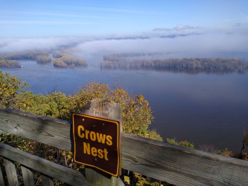 A scenic view of a river surrounded by fog and autumn trees, with a sign reading "Crows Nest" in the foreground.