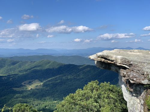 A rocky cliff overlooks a lush green valley and distant mountains under a blue sky with scattered clouds.