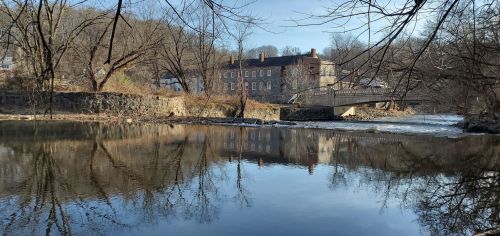 A serene river scene with a stone building and bridge reflected in the water, surrounded by bare trees and a clear sky.