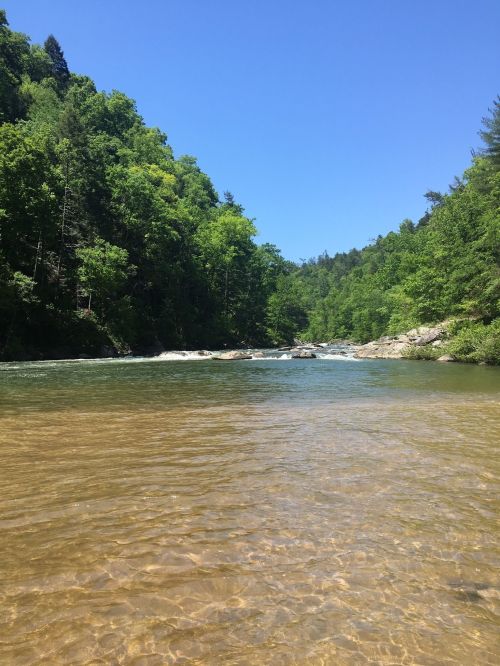 A serene river flows through lush green hills under a clear blue sky, with gentle ripples on the water's surface.