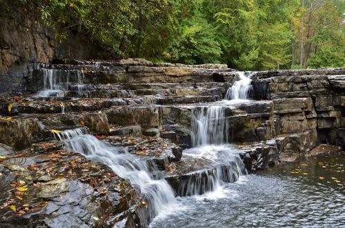 A serene waterfall cascading over rocky steps, surrounded by lush green trees and autumn leaves.