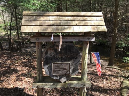 A wooden sign for a nature preserve, featuring a plaque and an American flag, surrounded by trees and a stream.