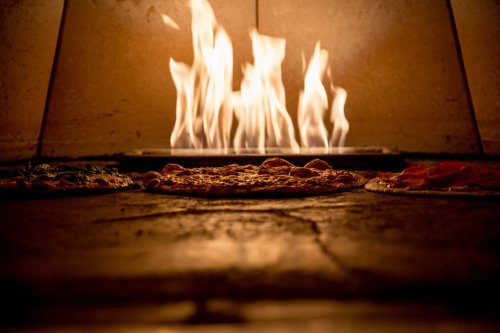 Three pizzas baking in a wood-fired oven, with flames flickering in the background.