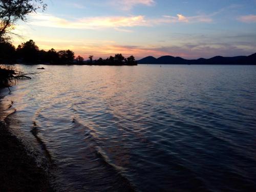 A serene lakeside view at sunset, with gentle waves and silhouettes of trees and mountains in the background.