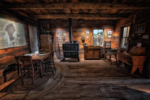 A rustic wooden interior featuring a dining table, stove, and various furnishings, illuminated by natural light.